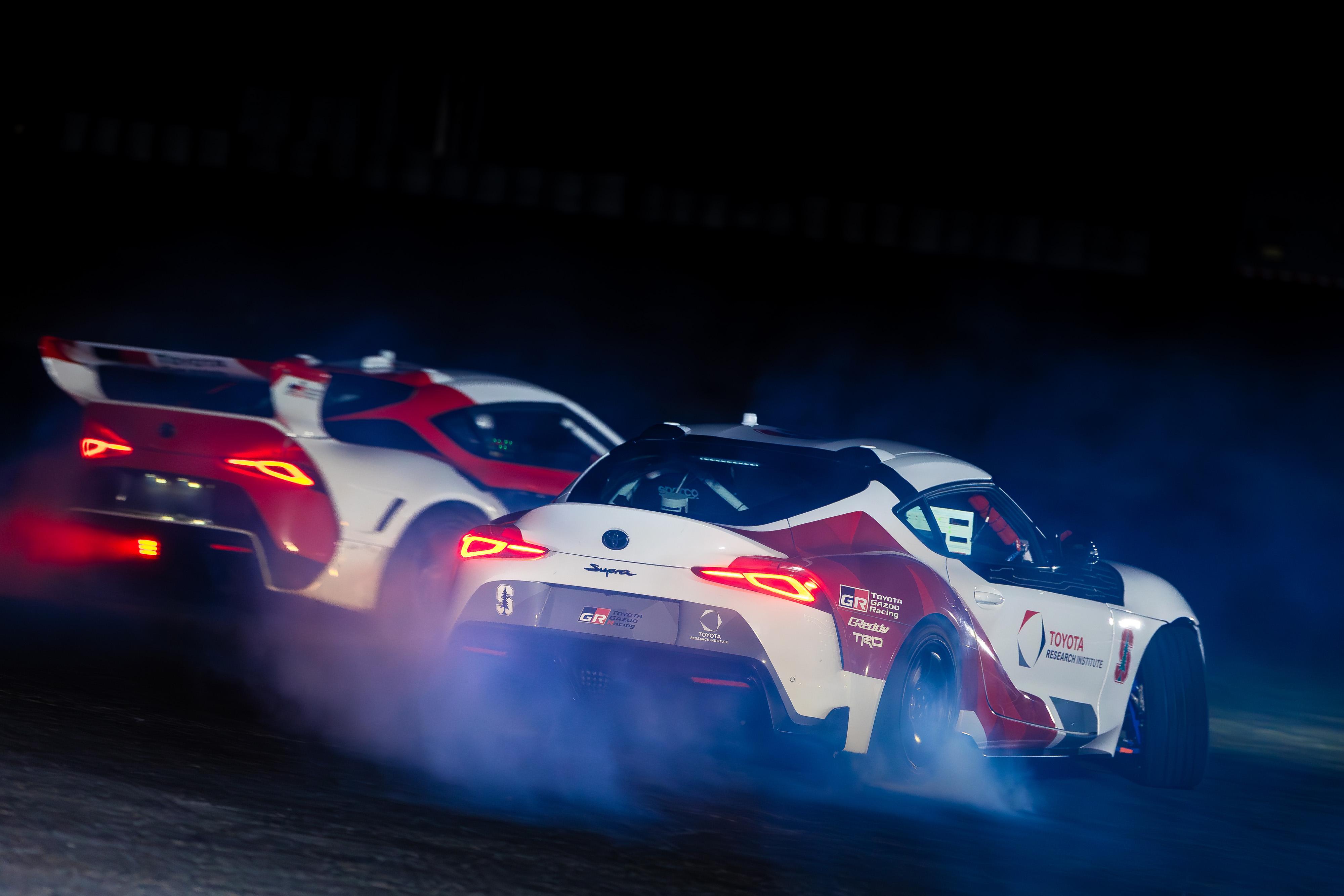 Smoke rises from the tires of two Toyota Supras tandem drifting autonomously